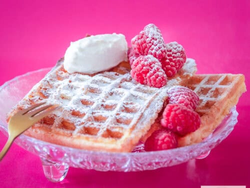 Een vierkante foto met een glazen schaaltje met 2 wafels, frambozen en mascarpone. Een roze achtergrond.