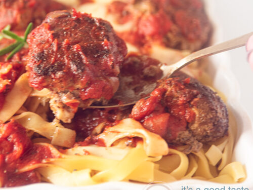 Een vierkante foto met een deel van een witte ovenschaal gevuld met tagliatelle, gehaktballejtes en tomatensaus. Een lepel houdt een balletje omhoog.
