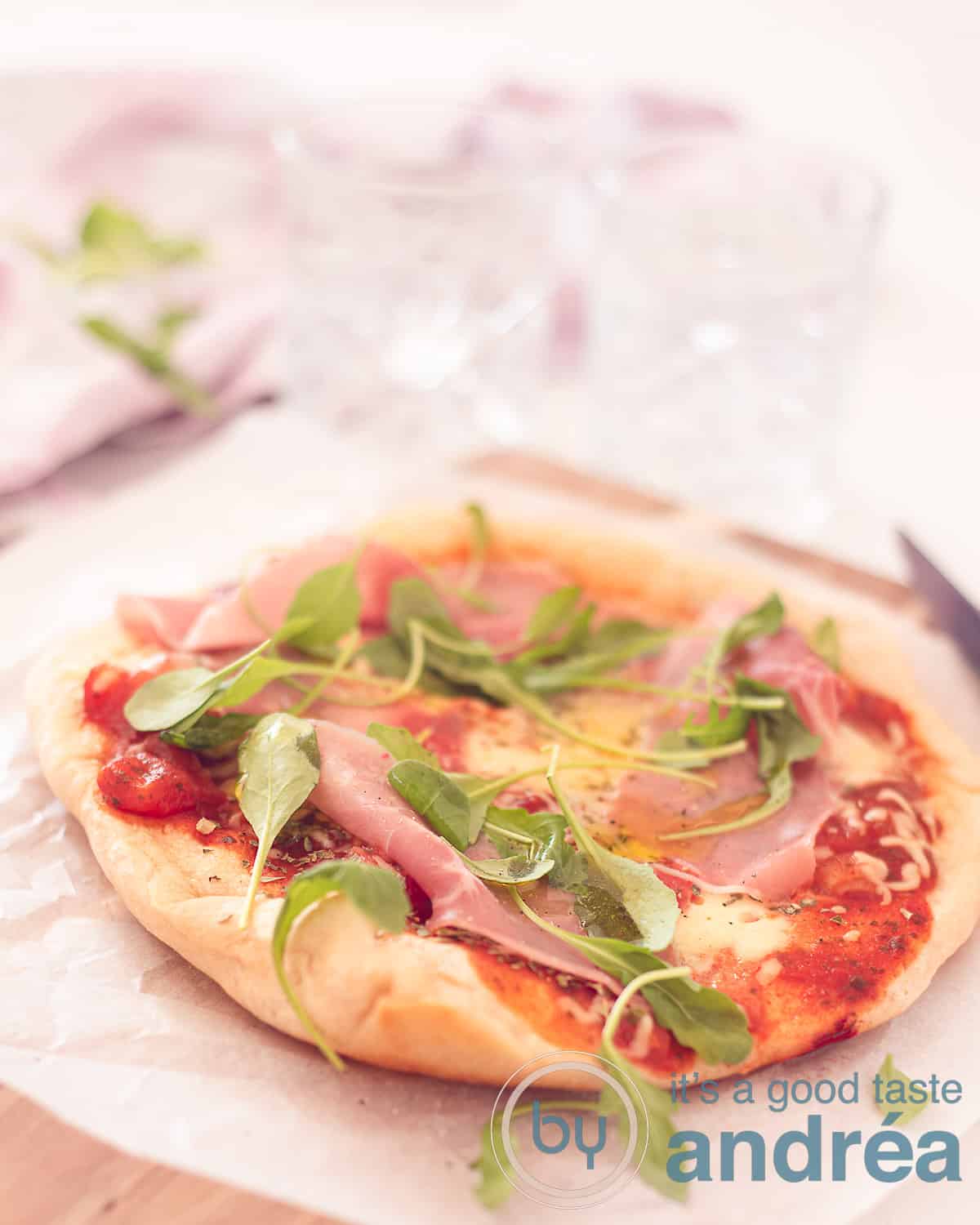 Een hoogtefoto met een pizza gemaakt in de koekenpan en belegt met tomatensaus, kaas, rucola en ham. Op een bakpapier op een houten plank.