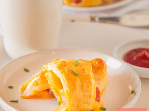 A square picture of a white plate with a cheesy pig in a blanket on top.