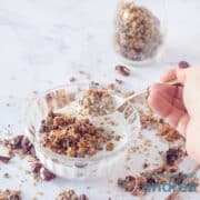 Een vierkante foto met een glazen schaaltje met yoghurt en chocolade muesli. Een gouden lepel neemt een hap erbij een marmeren witte ondergrond. In de achtergrond een glazen potje met meer chocolade noten granola.