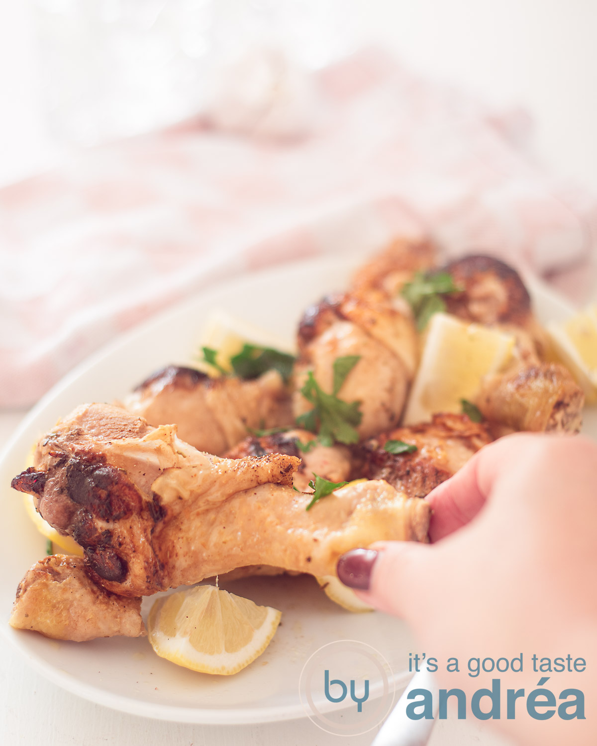 A white long plate with drumsticks, lemon parts and parsley. A hand taking a drumstick. A pink white cloth in the background