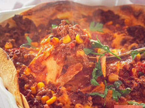 Een vierkante foto met een deel van een witte ovenschaal met gehakt, tomatensaus, tortilla's, mais en kaas.