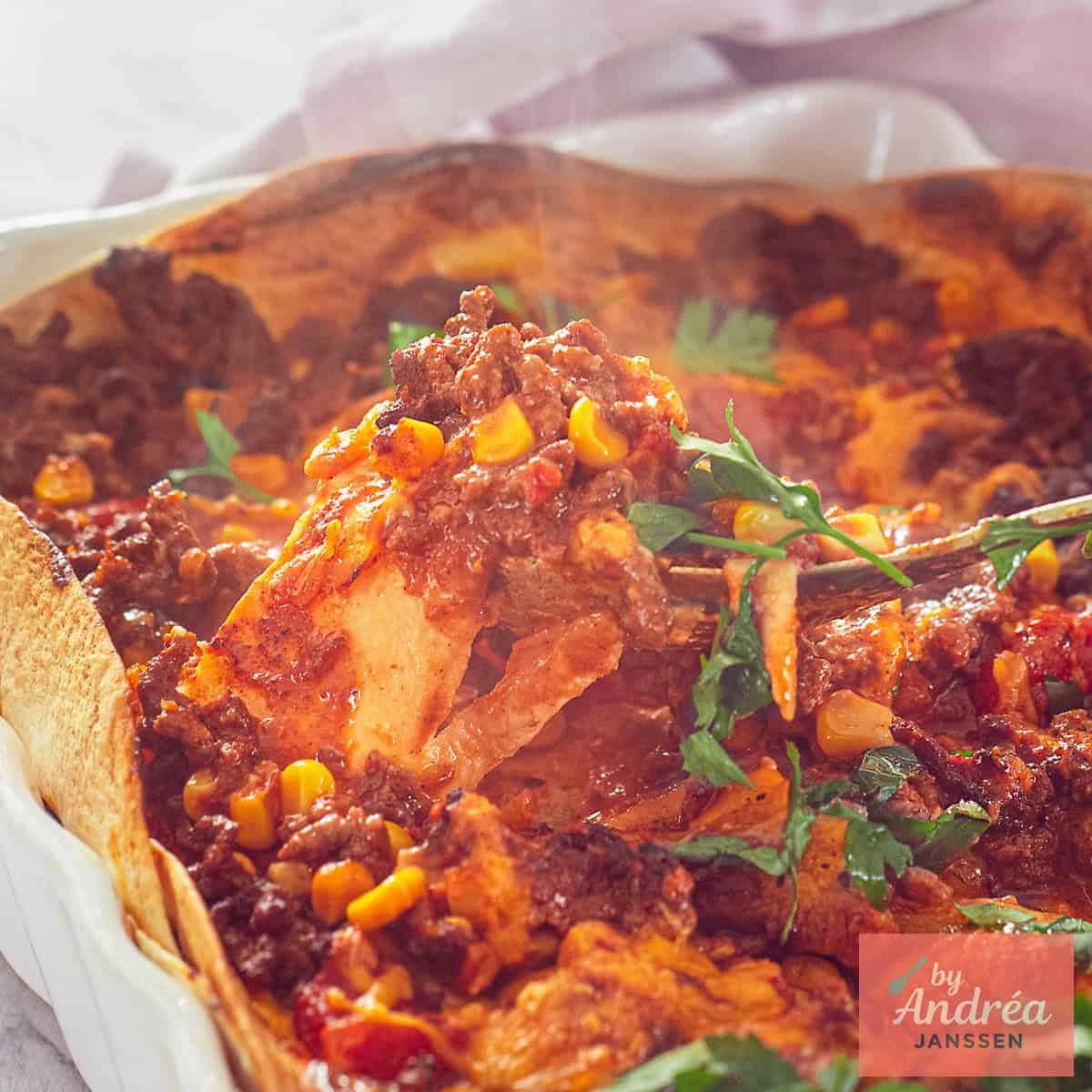 Een vierkante foto met een deel van een witte ovenschaal met gehakt, tomatensaus, tortilla's, mais en kaas.