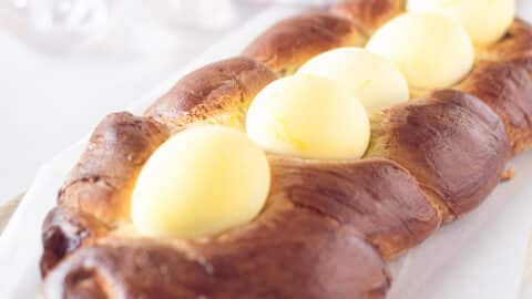 A square picture with Greek Easter bread topped with yellow eggs, on a white baking paper. A white background