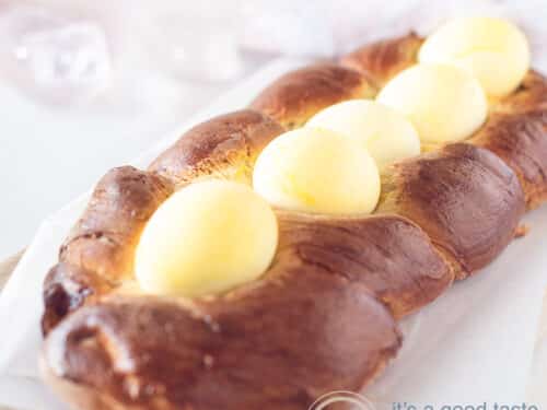 A square picture with Greek Easter bread topped with yellow eggs, on a white baking paper. A white background