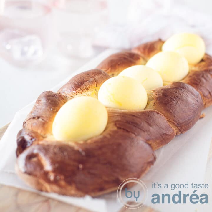 A square picture with Greek Easter bread topped with yellow eggs, on a white baking paper. A white background