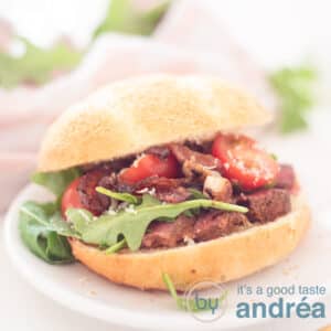 A square photo with an Italian bun, filled with slices of steak, caramelized onions, arugula, Parmesan cheese and tomatoes. A pink white cloth and rocket in the background,