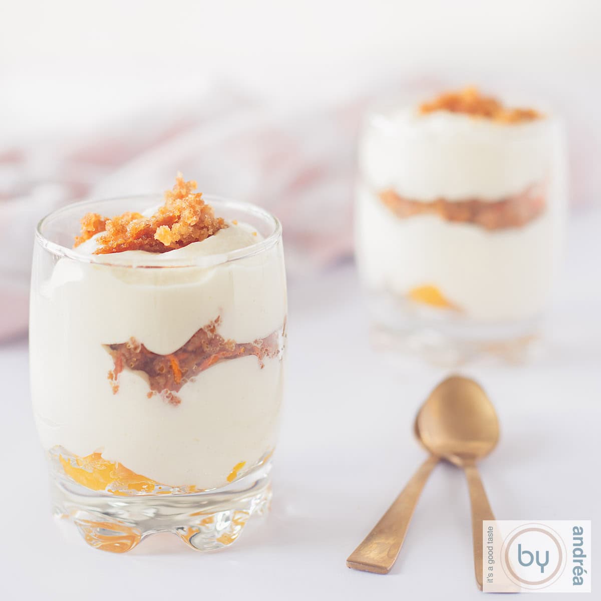 Een vierkante foto met een witte ondergrond met twee glaasjes gevuld met abrikozenjam, witte chocolademousse en wortelcake. Twee lepels links van de glaasjes