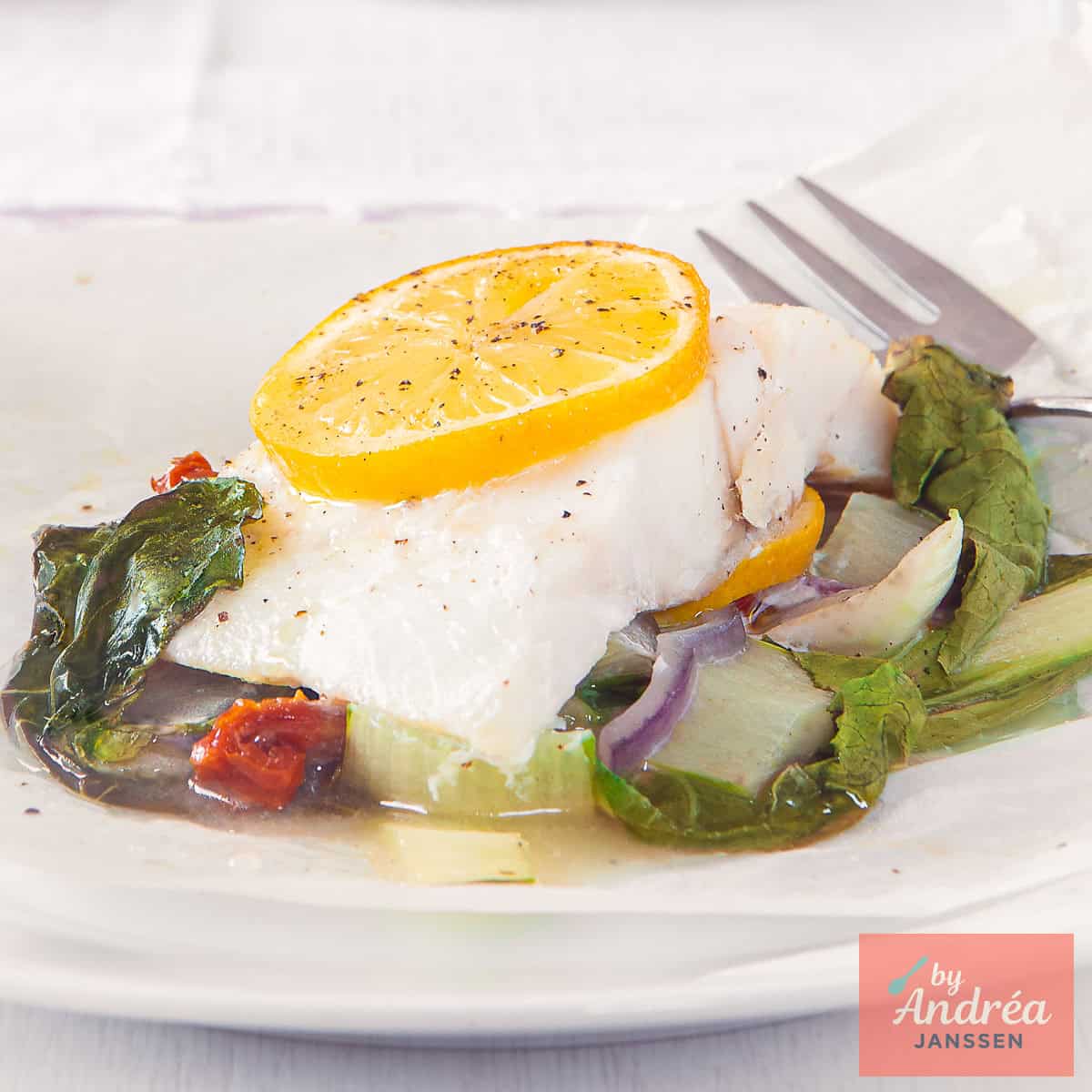 Een vierkante foto met een bord met paksoi, zongedroogde tomaten en rode ui met daarop vis met citroen.