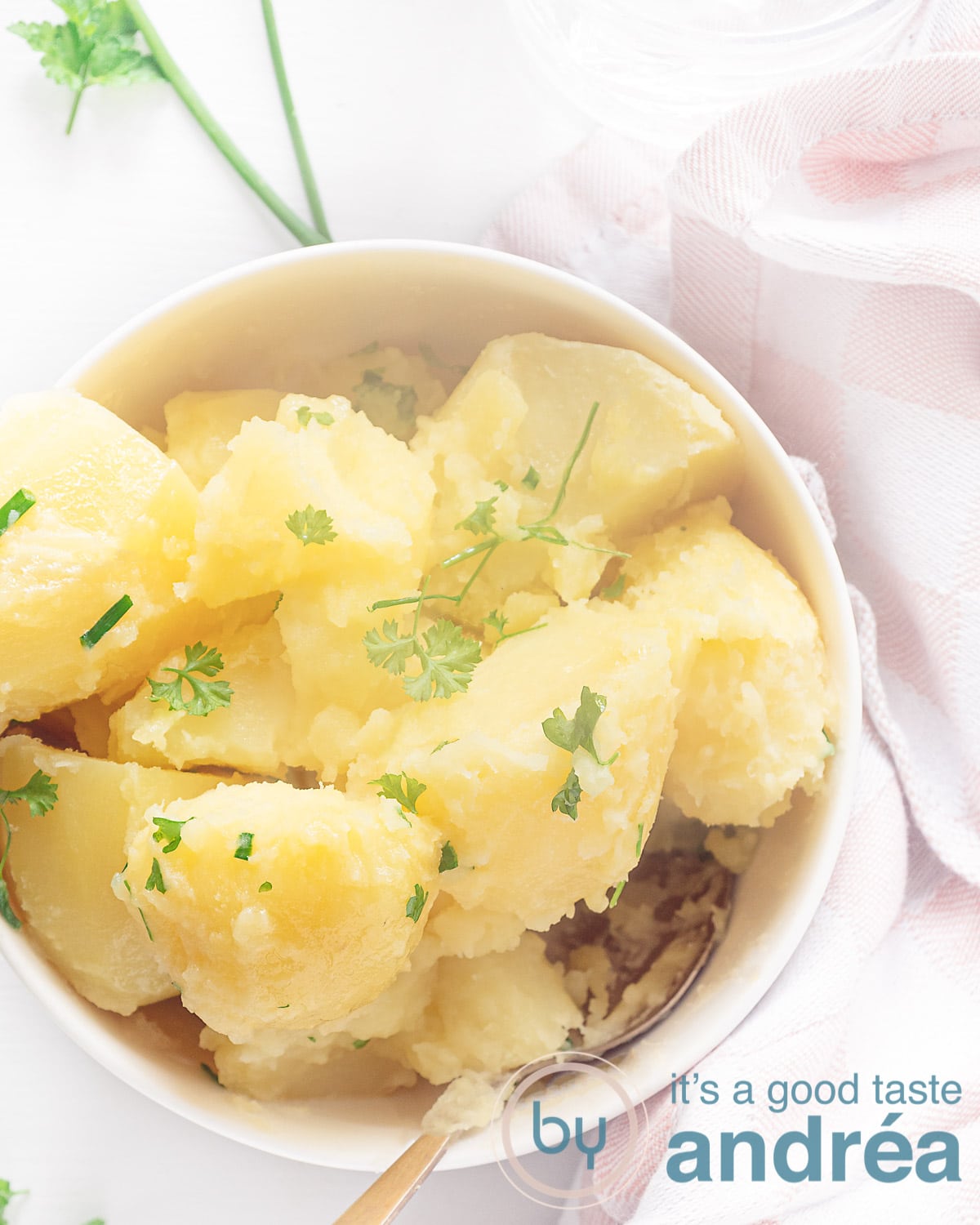 Een foto van bovenaf van een witte schaal gevuld met aardappels die gekookt zijn met bieslook en peterselie. Een witte ondergrond met deel van een roze witte theedoek