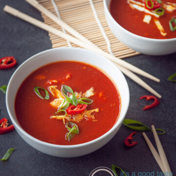 Twee kommen gevuld met chinesetomatensoep en een garnering van ei reepjes, lente ui, en peper. Een zwarte ondergrond. Tussen de kommen liggen chopsticks.De achterste kom staat half op een rieten matje.