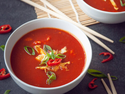 Twee kommen gevuld met chinesetomatensoep en een garnering van ei reepjes, lente ui, en peper. Een zwarte ondergrond. Tussen de kommen liggen chopsticks.De achterste kom staat half op een rieten matje.