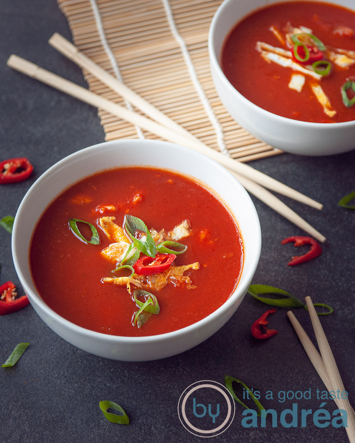 Twee kommen gevuld met chinesetomatensoep en een garnering van ei reepjes, lente ui, en peper. Een zwarte ondergrond. Tussen de kommen liggen chopsticks.De achterste kom staat half op een rieten matje.