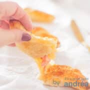 Een vierkante foto met drie ham-kaas croissants achter elkaar. De eerste croissant is opengebroken en een hand houdt een stuk vast. Er tussen zie je gesmolten kaas en ham.
