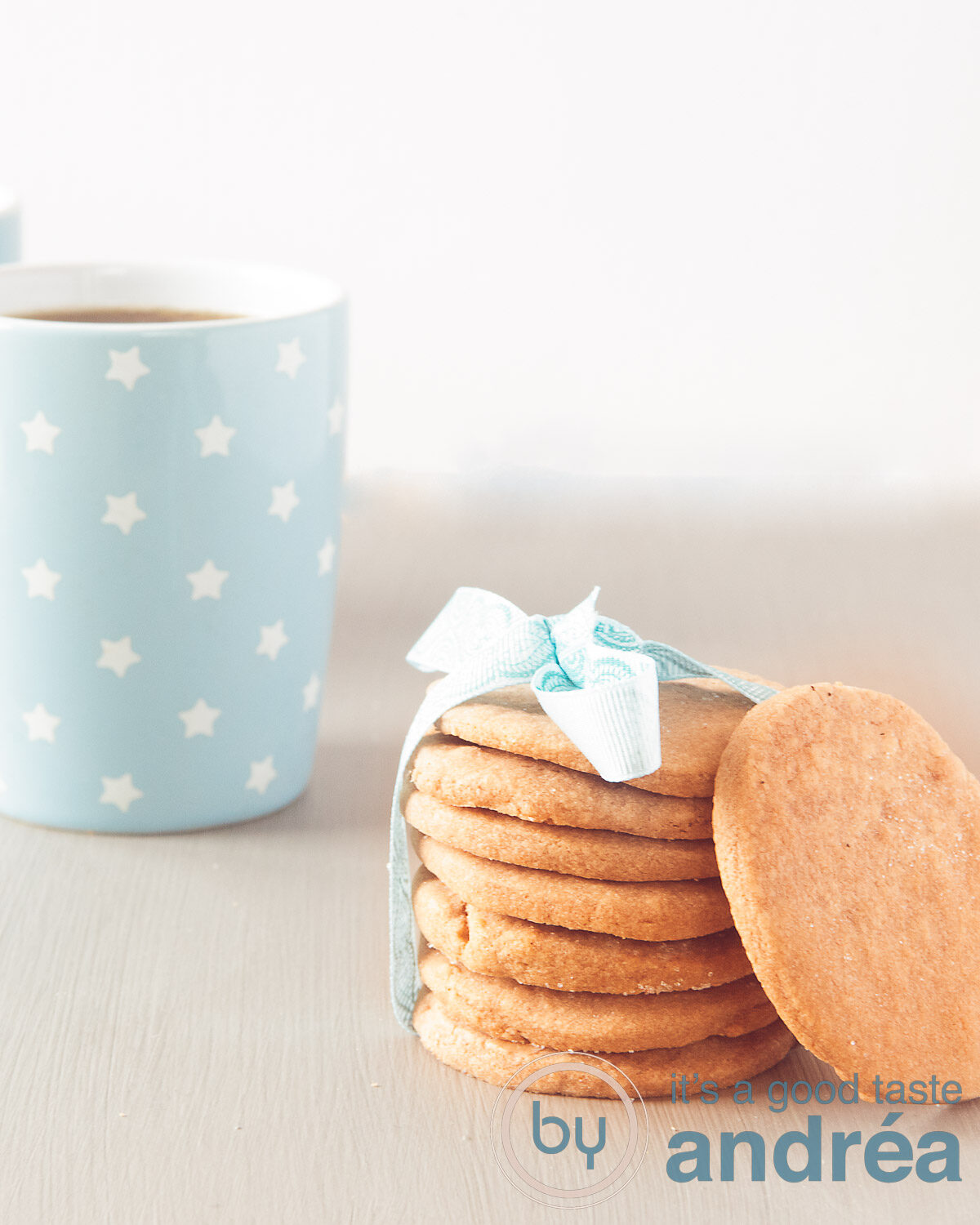 Een grijze ondergrond met een stapel koekjes met een blauw lintje een koekje ertegenaan. Een blauw kopje koffie er schuin achter