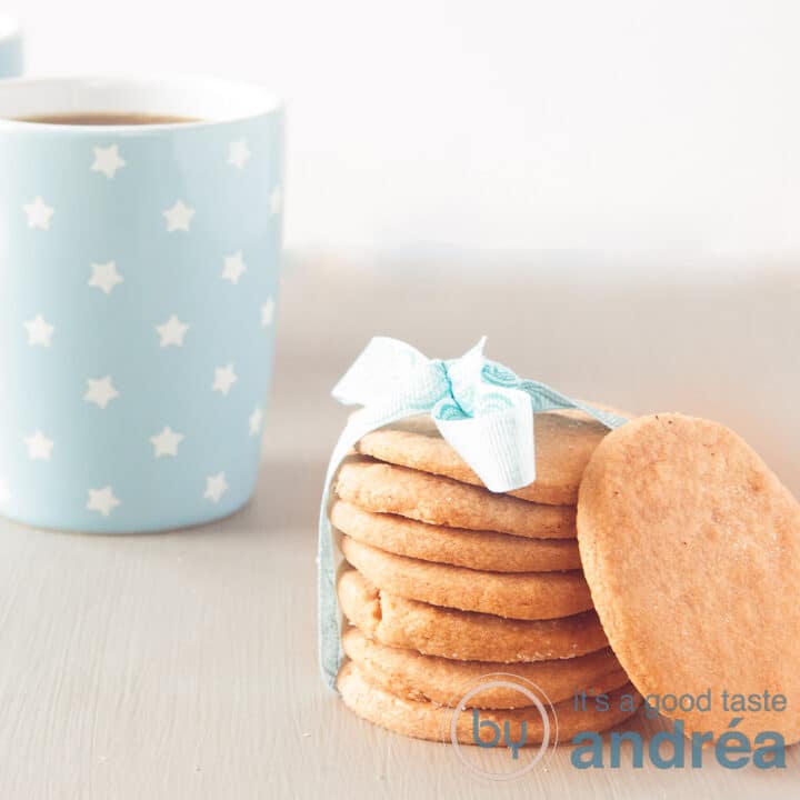 Een vierkante foto van een grijze ondergrond met zeven gestapelde koekjes met lint eromheen. Een koekje staat er tegenaan. In de achtergrond een mok met koffie