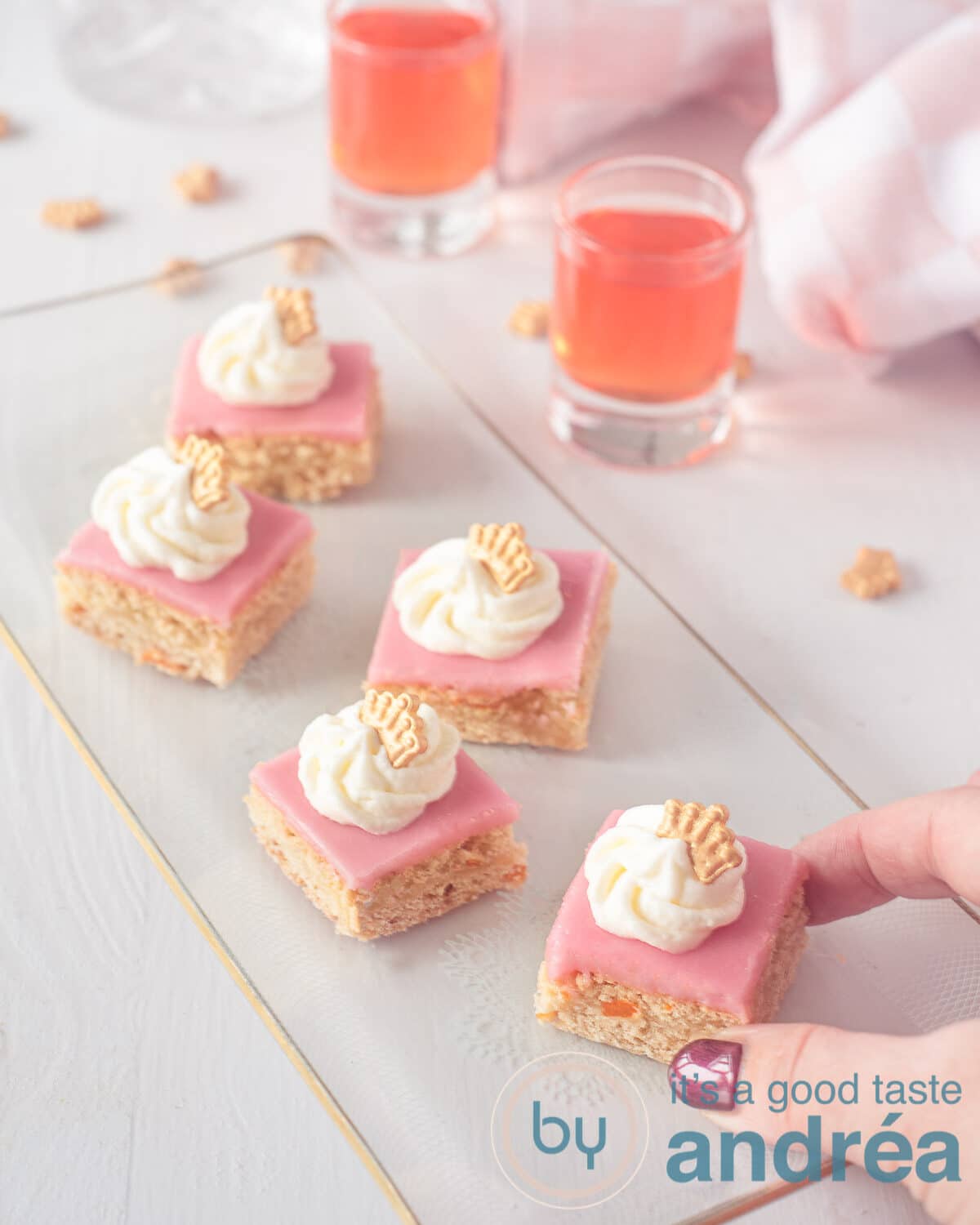 Een cakeschaal vol stukjes Oranje koek. Een hand pakt een blokje. Een witte ondergrond. Op de achtergrond twee oranjebitters.