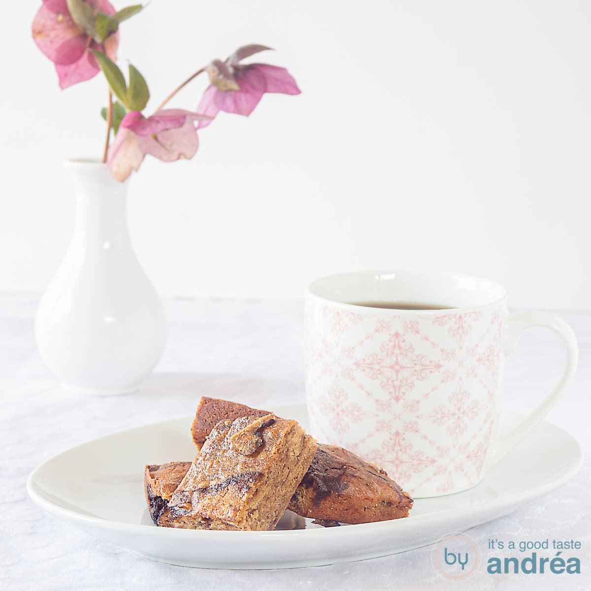 Een vierkante foto van een wit gebaksbordje met een paar stukjes blondie met honing en chocola. Een roze wit theekopje op het schaaltje. In de achtergrond een vaasje met bloemen.