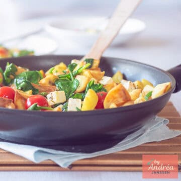 Een vierkante foto met een deel van een wok gevuld met geroerbakte aardappeltjes, feta, spinazie en tomaat.