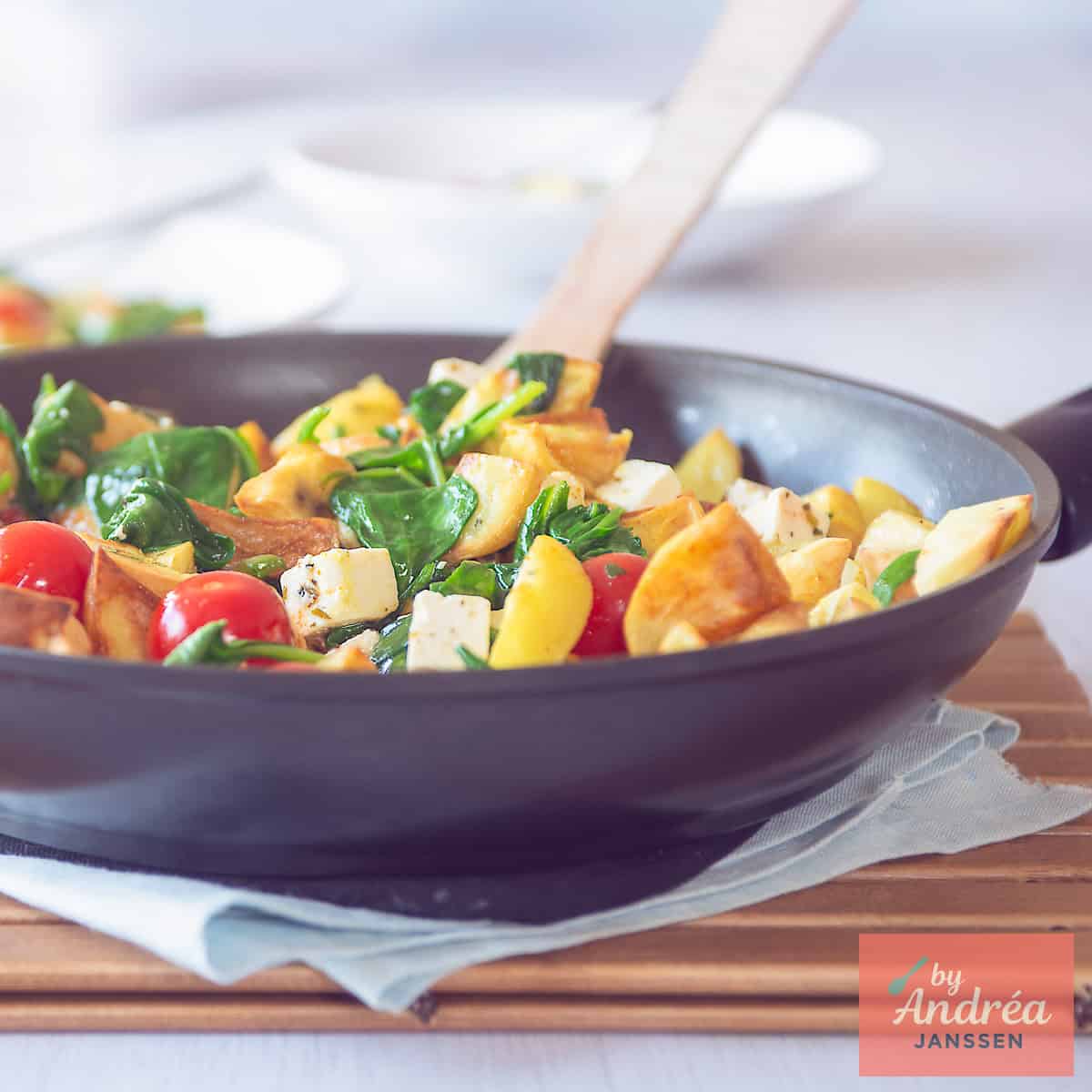 Een vierkante foto met een deel van een wok gevuld met geroerbakte aardappeltjes, feta, spinazie en tomaat.