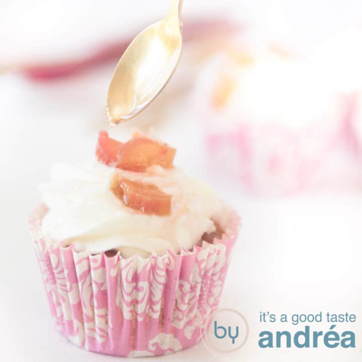 Een vierkante foto van een licht roze cupcake vorm met een slagroom rabarber topping. Van een lepel sprenkelt wat siroop.