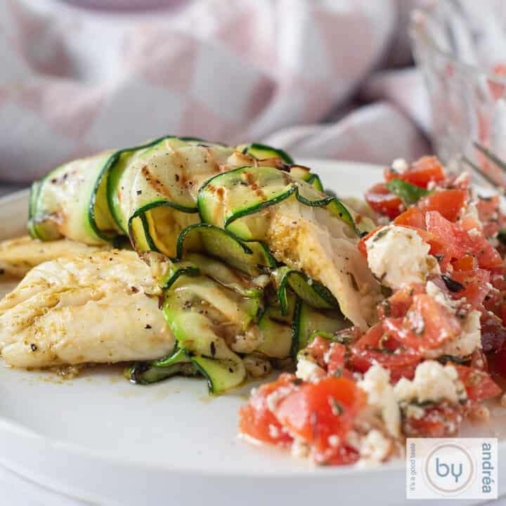 Een vierkante foto met een wit bord waarop tilapia filet gerold in courgette met een heerlijke tomatensalade ligt.