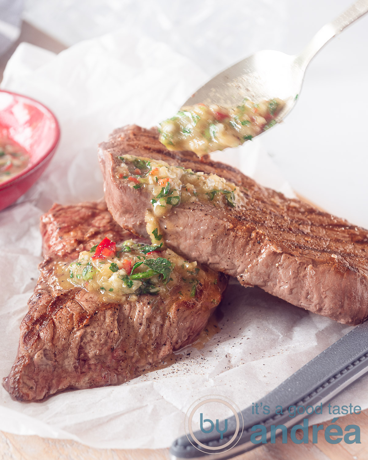 Een hoogtefoto waar twee stukken steak op een bakpapier liggen en een gouden lepel chimichurri saus erop schenkt
