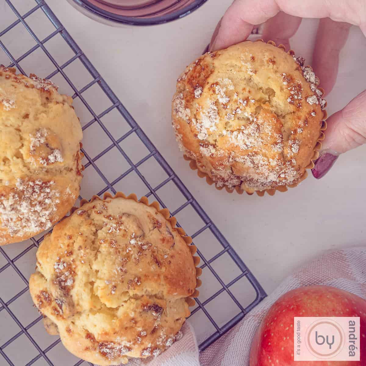 Een vierkante foto van drie appelmuffins met rozijnen. Een hand pakt er een.