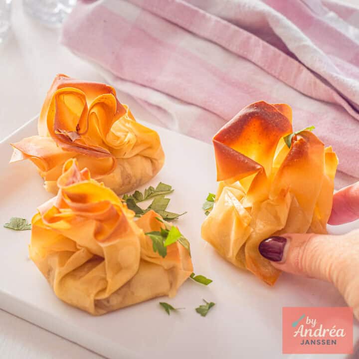 A square picture with a white plate with three  Goat Cheese Phyllo Appetizers. A hand taking one.