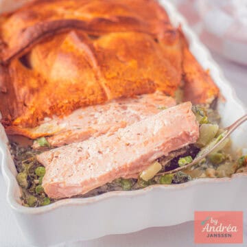 Een vierkante foto met een ovenschotel met groene groenten, zalm en een laagje croissantdeeg.
