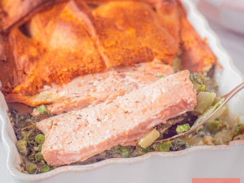 Een vierkante foto met een ovenschotel met groene groenten, zalm en een laagje croissantdeeg.