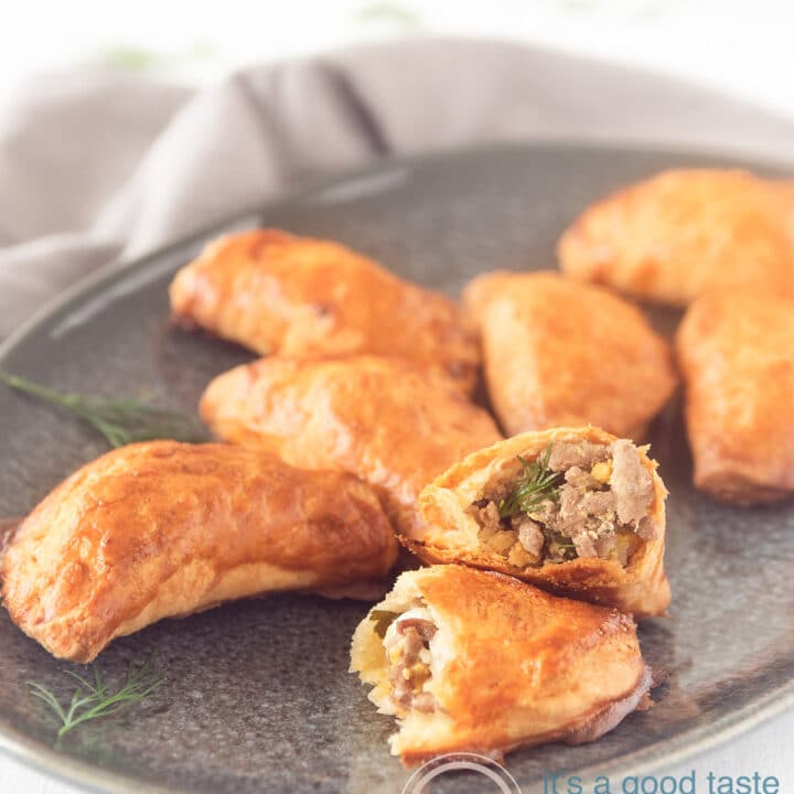 Russische pirozhki op een grijze schaal. Eén snack in tweeën gebroken. Dille is ter decoratie eromheen gespreid.