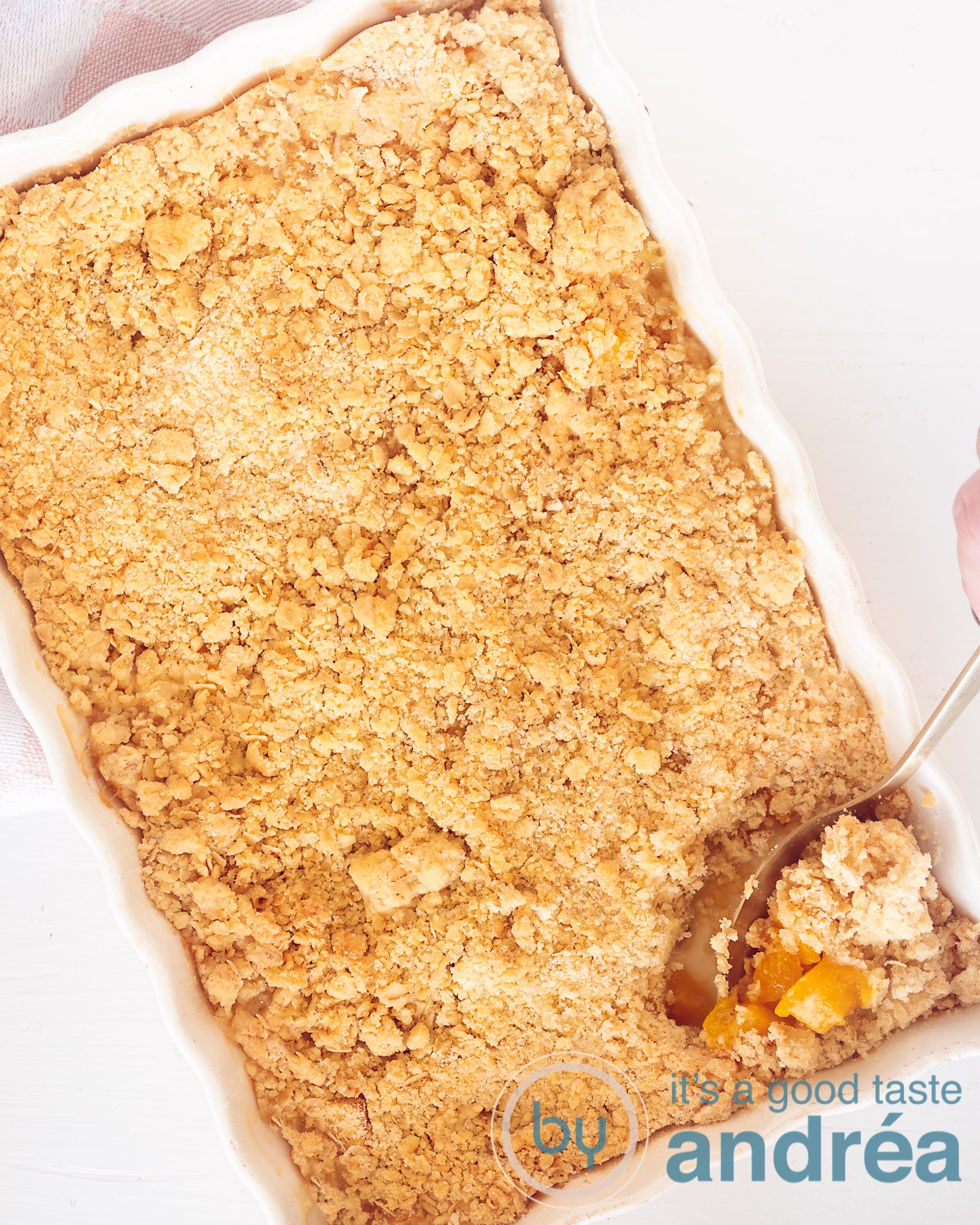 Een foto van bovenaf met een witte ovenschaal met een crumble, rechtsonderin een lepel eruit