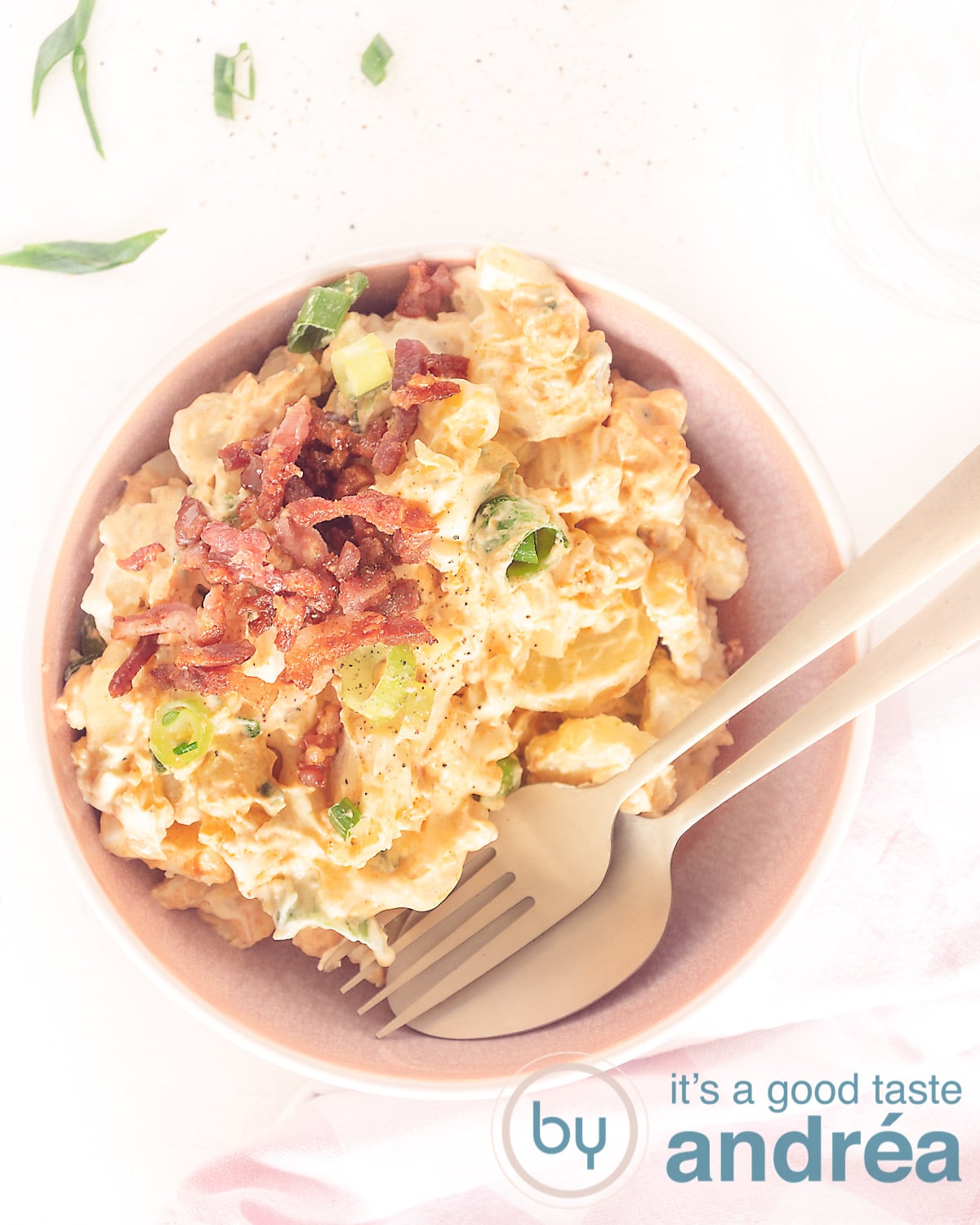 Een foto van bovenaf van een roze schaal gevuld met aardappelsalade, groene lente uites en bacon