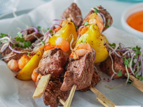 Een vierkante foto met een witte ondergrond met vijf spiesjes surf en turf biefstuk met garnalen. Een bakje met chilisaus en een blauwe doek in de achtergrond.