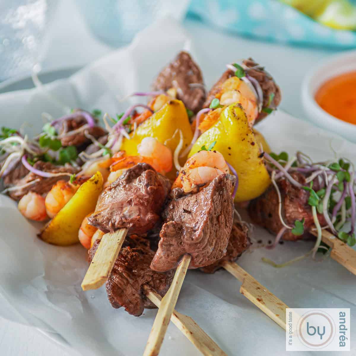 Een vierkante foto met een witte ondergrond met vijf spiesjes surf en turf biefstuk met garnalen. Een bakje met chilisaus en een blauwe doek in de achtergrond.