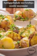 Een beige schaal met een aardappelsalade van krieltjes. Een lepel schept wat aardappels eruit. Een tekst bovenin: aardappelsalade met krieltjes zonder mayonaise.
