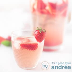 Een vierkante foto met voorin een glas aardbeienlimonade met verse aardbeien en achterin een kan limonade