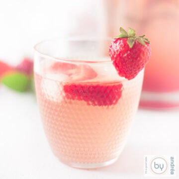 Een vierkante foto met een glas aardbeienlimonade met ijs en verse aardbeien in de voorgrond.