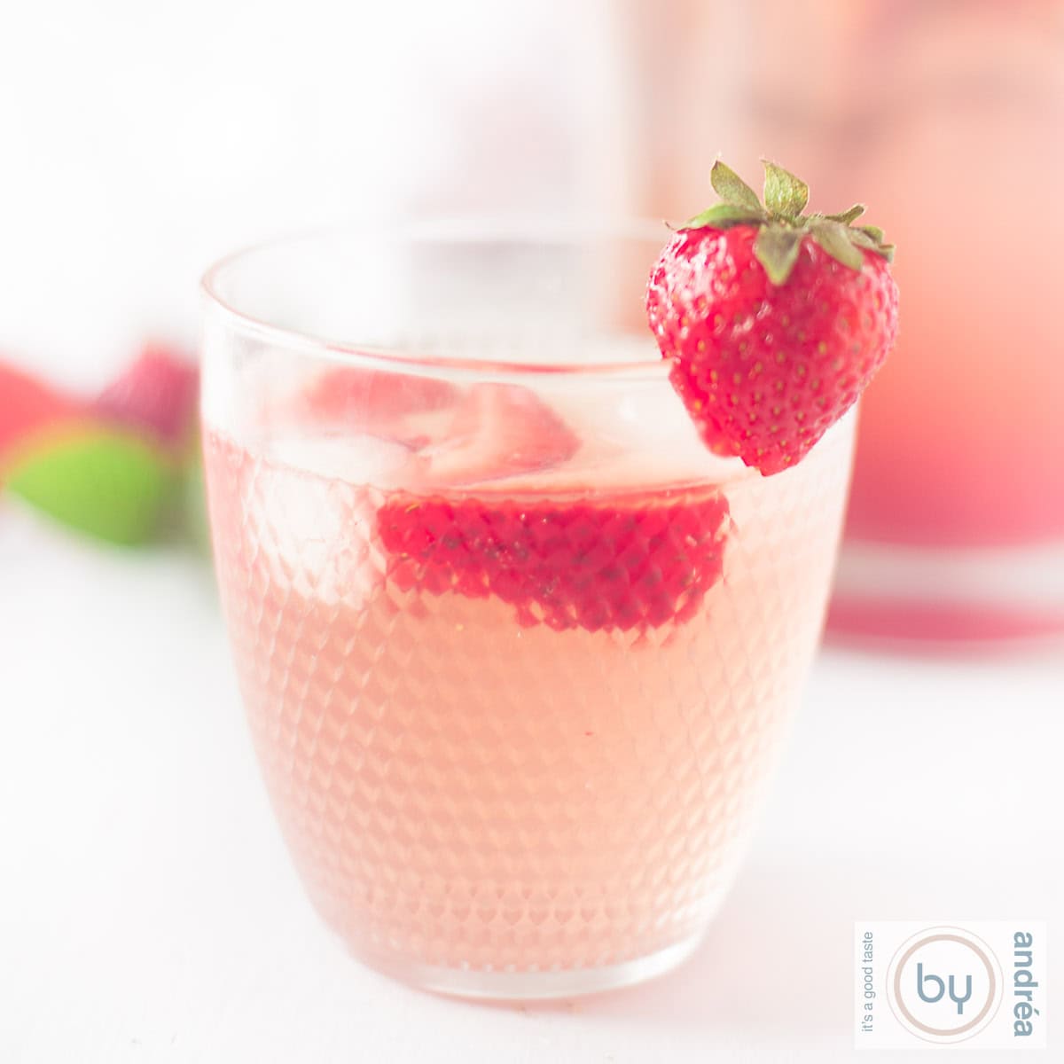 Een vierkante foto met een glas aardbeienlimonade met ijs en verse aardbeien in de voorgrond.