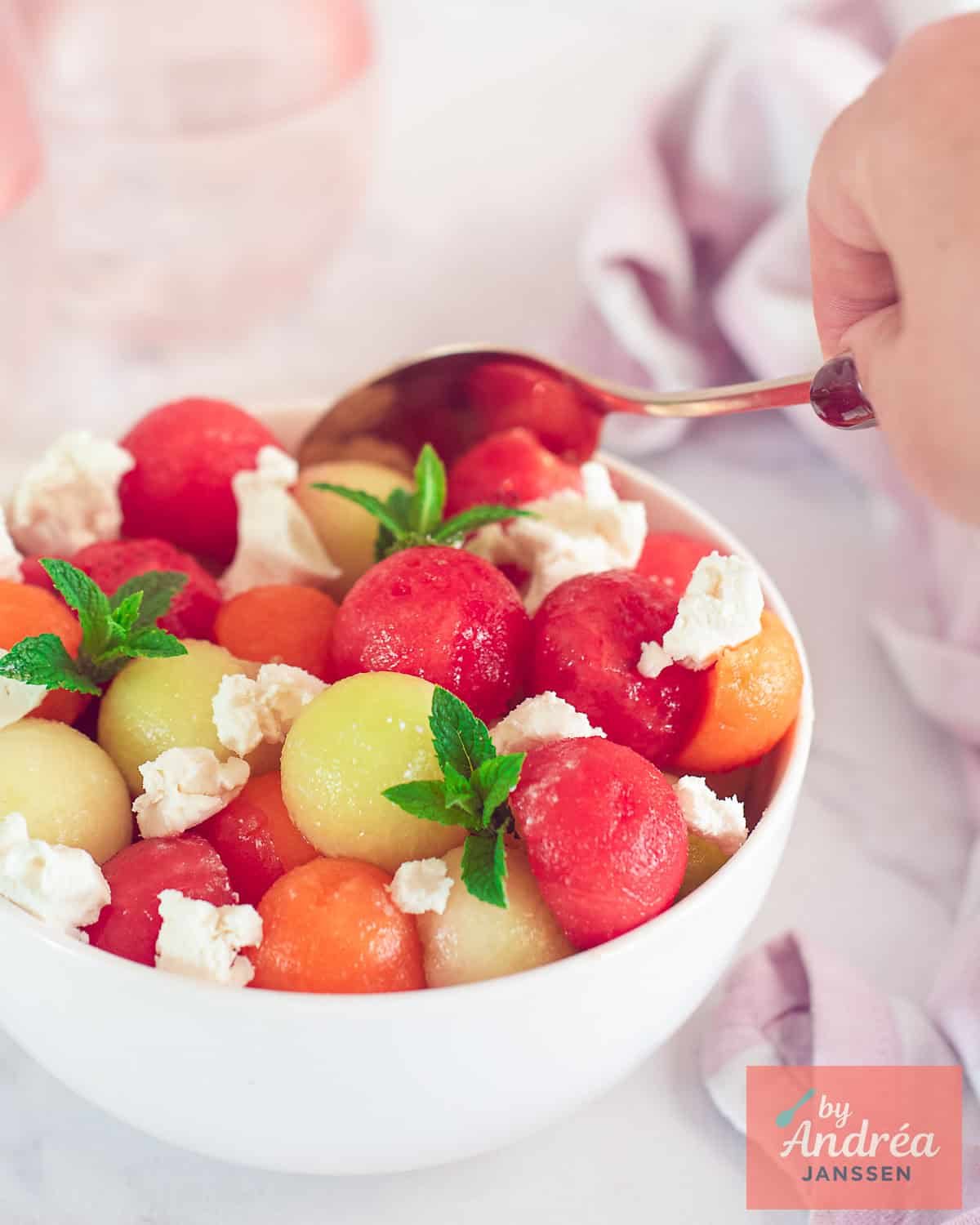 A portion of a white bowl with three-colored melon balls, mint, and goat cheese. A spoon takes a scoop