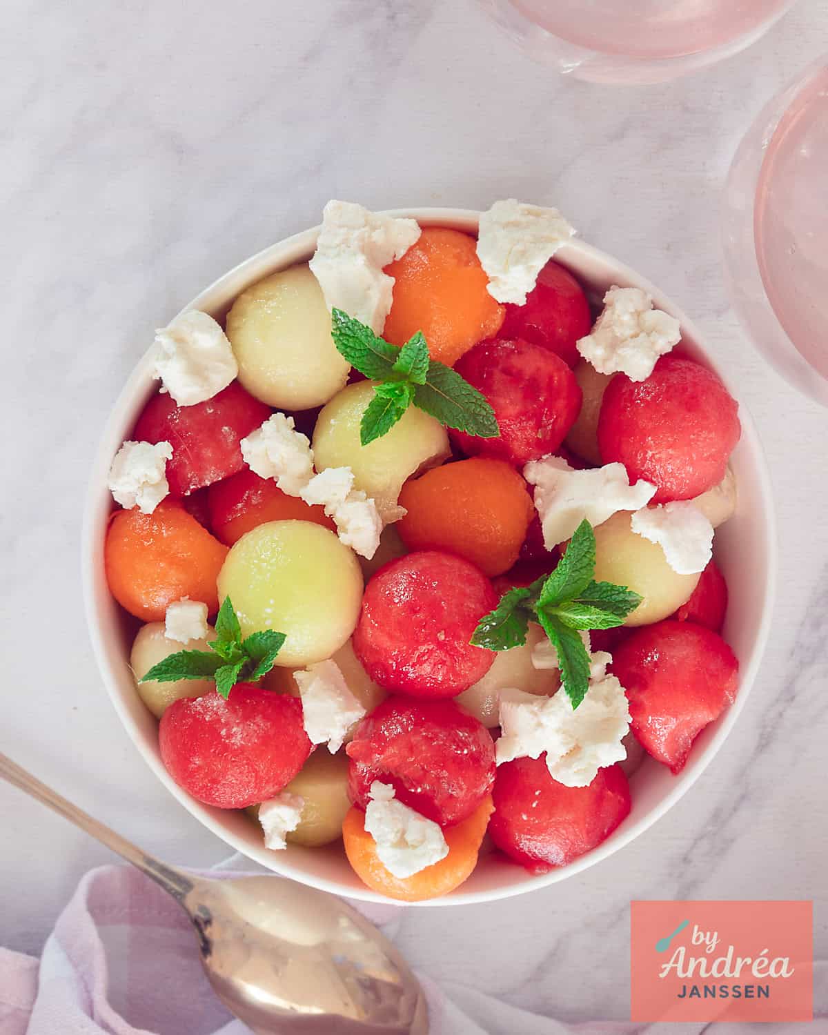 An overhead shot of a bowl of three-colored melon balls (galia, watermelon, and cantaloupe) with goat cheese, honey, and mint.