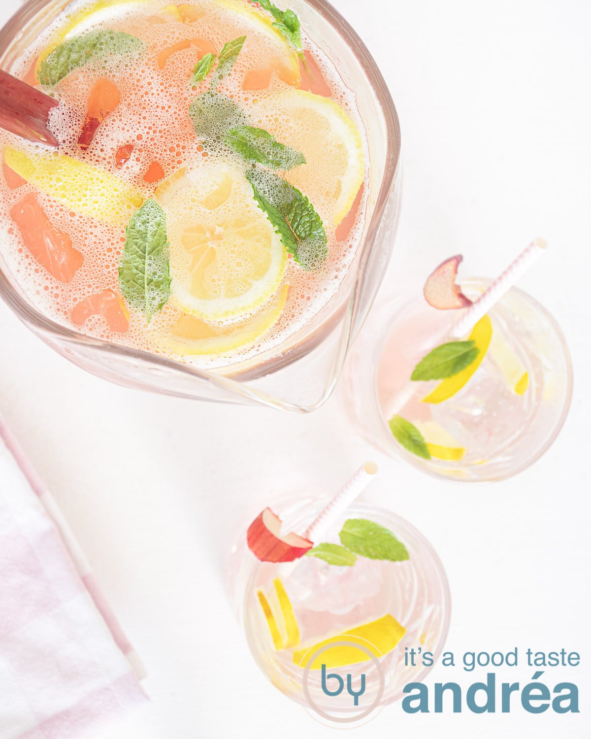 Een foto van bovenaf van een grote kan met citroen, munt en rabarber. Twee glazen gevuld met ijthee op de voorgrond. Een witte ondergrond