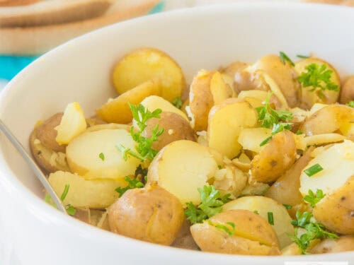 Een vierkante foto met een deel van een witte schaal met gehalveerde krieltjes, kruiden in een heerlijke salade dressing.