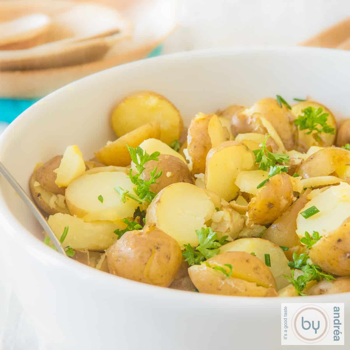 Een vierkante foto met een deel van een witte schaal met gehalveerde krieltjes, kruiden in een heerlijke salade dressing.