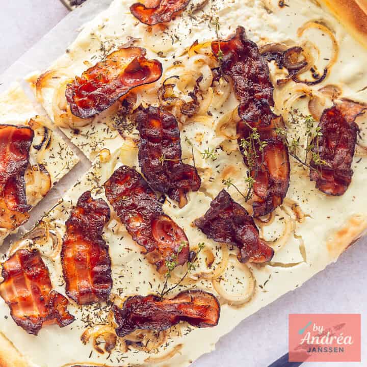 Een vierkante foto met een deel van een flamkuchen met crème fraîche topping, spek, tijm en uien.