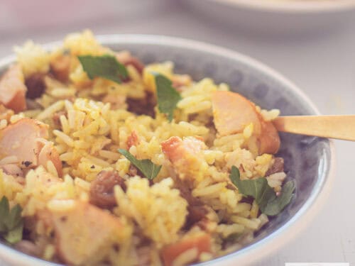 Een vierkante foto met een grijs schaaltje gevuld met een rijstsalade met kerrie, gerookte kip, rozijnen en walnoten