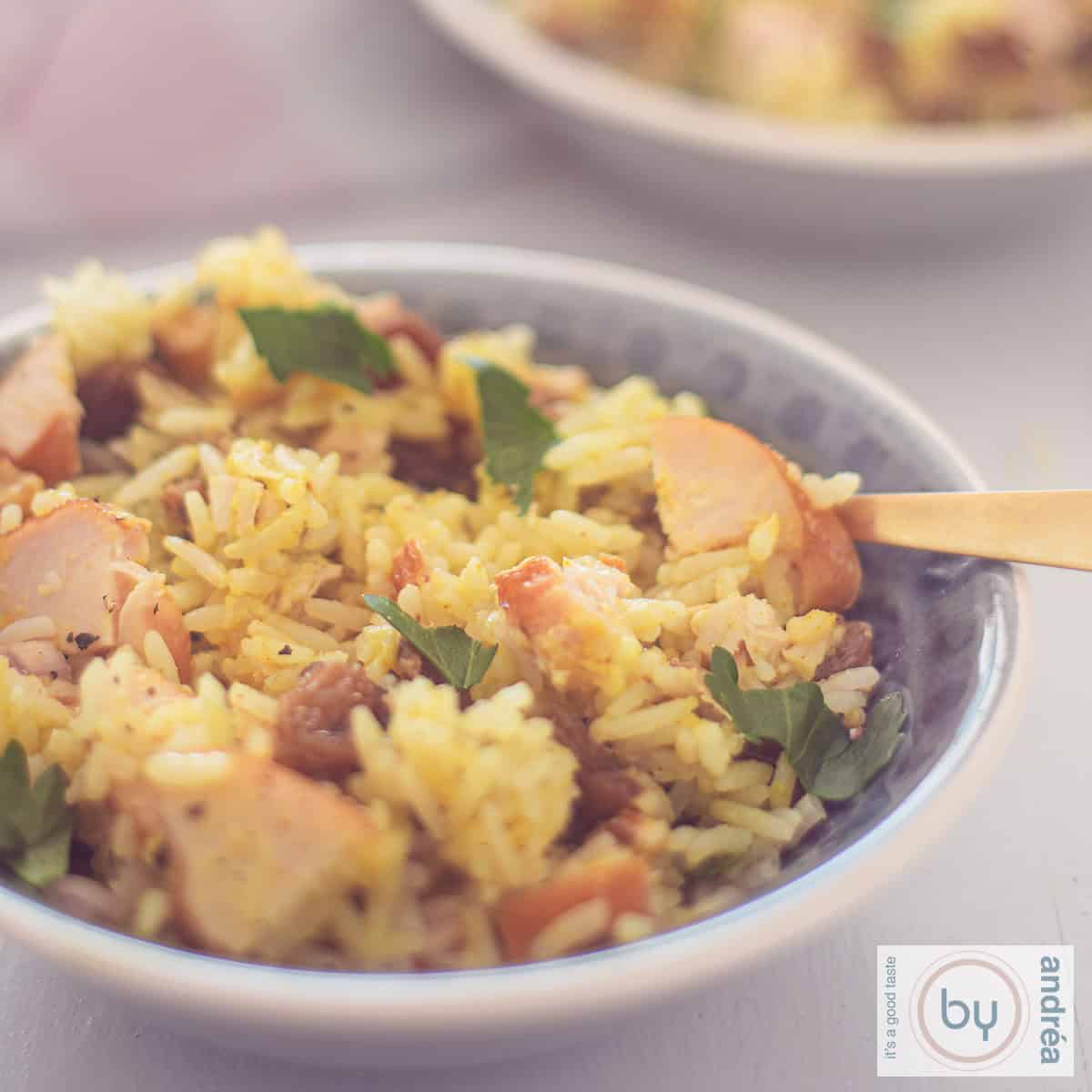 Een vierkante foto met een grijs schaaltje gevuld met een rijstsalade met kerrie, gerookte kip, rozijnen en walnoten