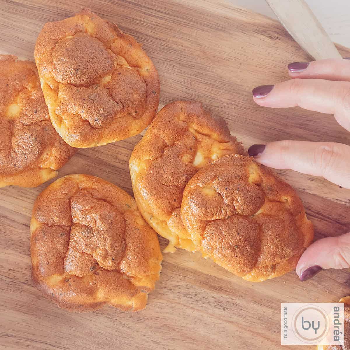 Een vierkante foto met een houten plank met daarop cloud breads. Een hand pakt een oopsie